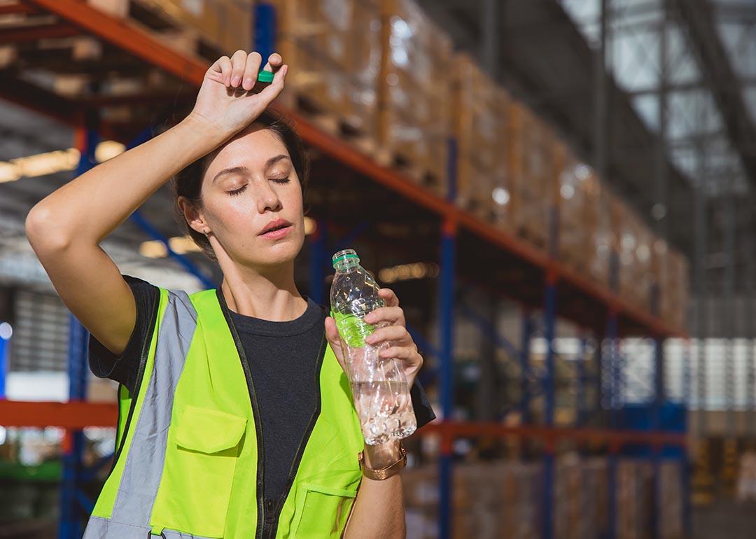 Woman wearing a high visibility vest holds a bottle of water and brushes off her forehead while her eyes are closed; stacks of pallets in a warehouse in the background.