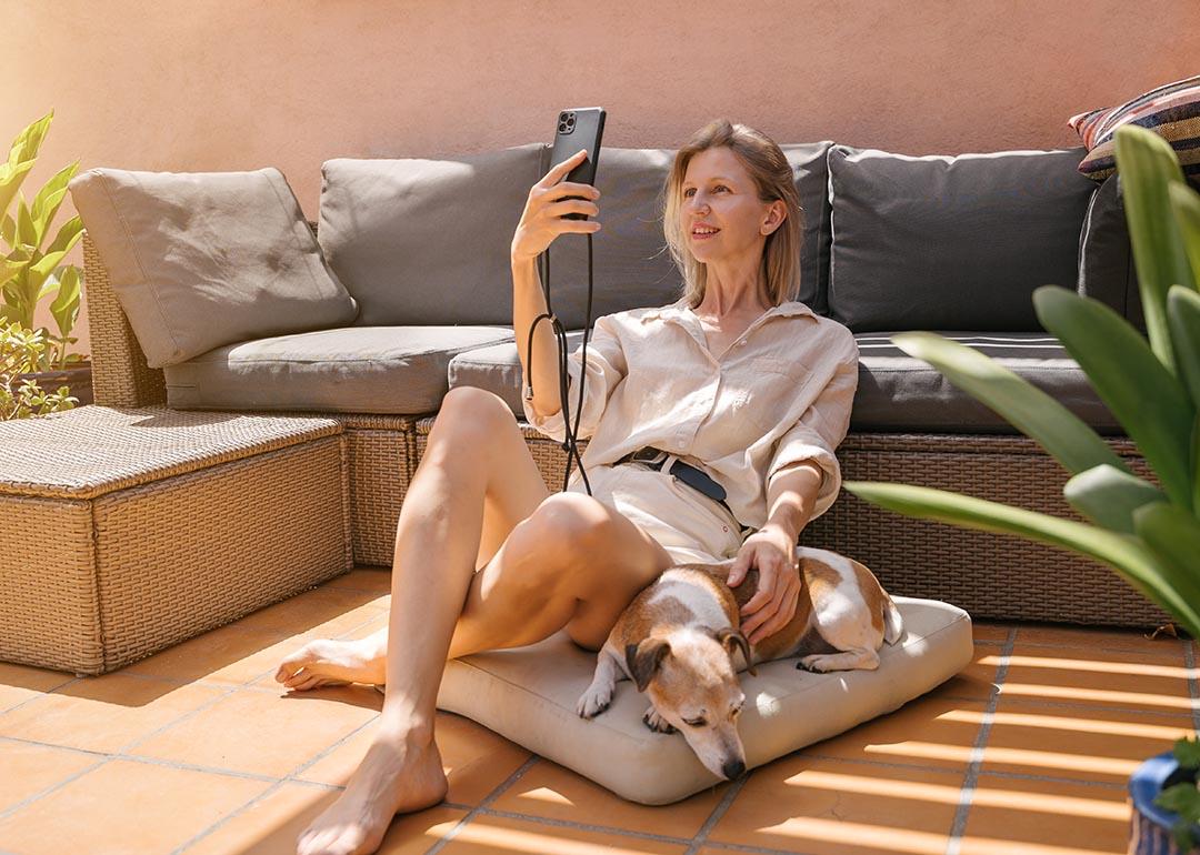 Woman wearing neutral button up top and shorts sitting on a floor cushion leaning against a couch, next to her dog, facetiming on her phone.