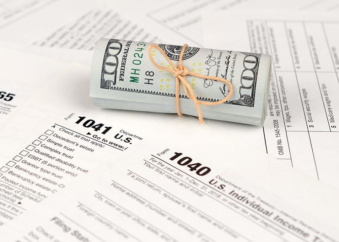 A tied up roll of hundred dollar bills on top of various paper U.S. tax forms like the 1040 and 1041.