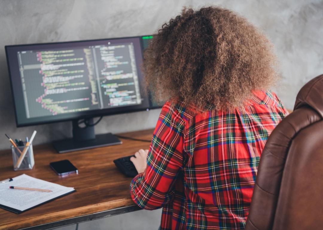 Rear view of a woman with curly hair working at a computer desk displaying code in a casual office setup wearing a checked shirt.