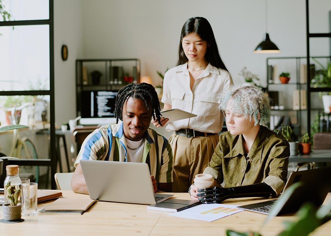 Three diverse young adults gathered around a laptop in a modern office setting.