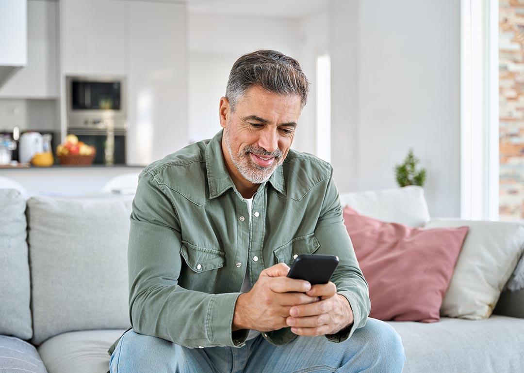 Middle aged old man using smartphone sitting on a couch with kitchen in background.