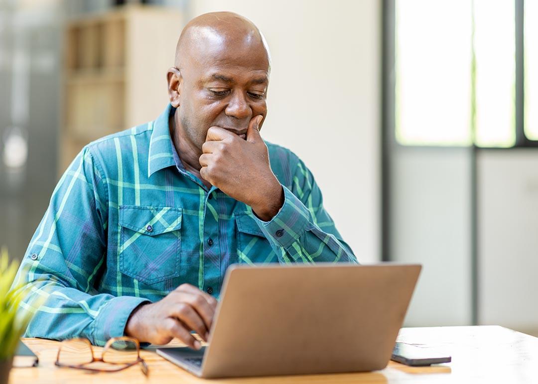 Black bald man wearing teal button up shirt using laptop computer and thinking about question in office.