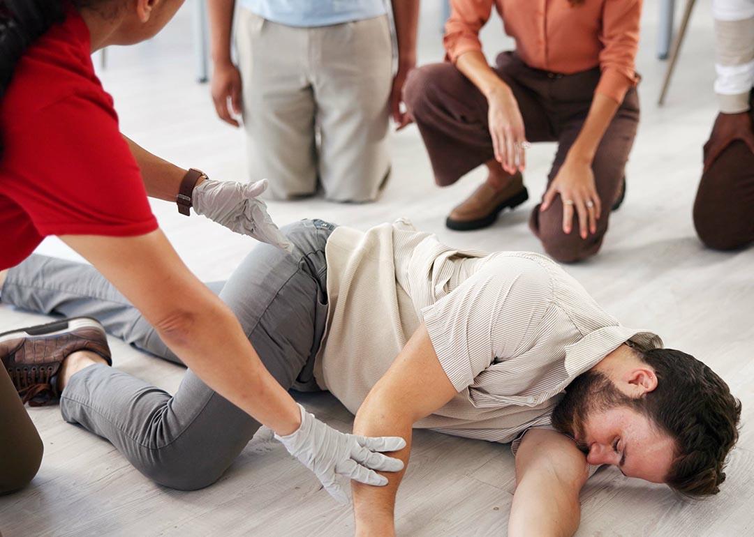 Demonstration by person teaching class for first aid, with someone as victim lying on their side and three people kneeling and watching.