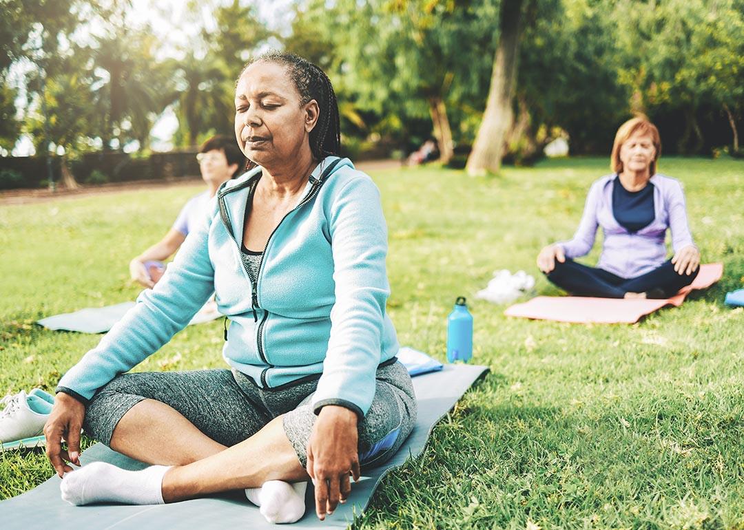 Three people in seated yoga poses on individual yoga mats on a green lawn, not sitting close together.
