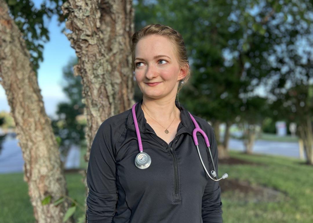 Michaela Bonner, wearing a black zip-up top with a pink stethoscope around her neck, poses for a portrait in front of some trees at local hospital in Virginia Beach, VA.