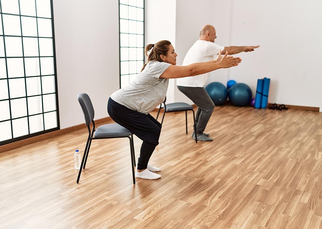 Two people each squatting toward a chair with their arms out in front of them, in a yoga studio type room.
