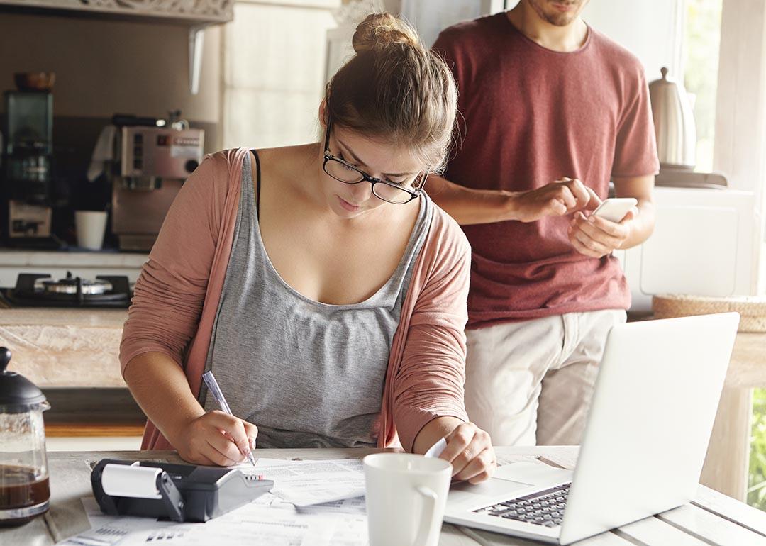 Woman doing calculations at kitchen table with laptop open and pen and paper while man with face not visible in the background is on his phone.