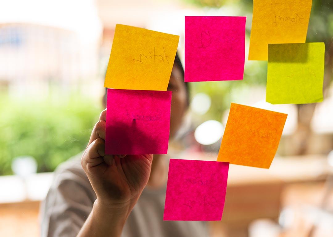 7 Pink, orange and yellow post-it notes stuck on glass with person whose face is obscured, writing on them from the other side.