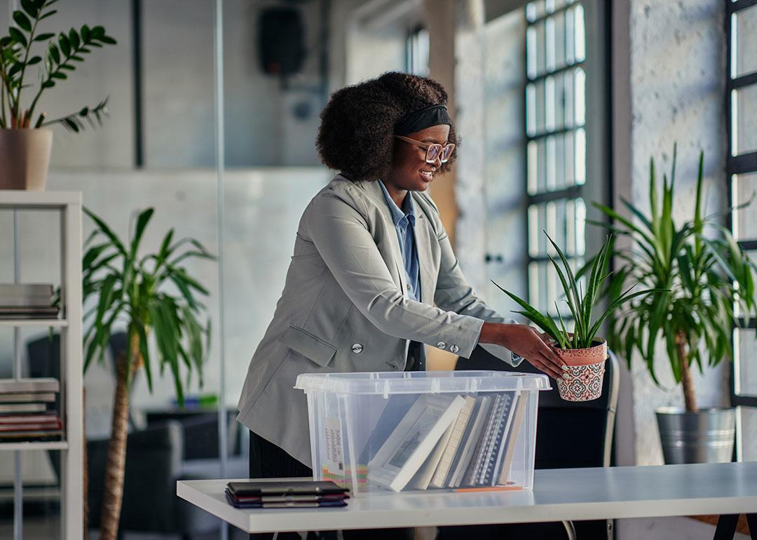 Black woman with glasses placing plants on an office desk with a clear plastic bin full of belongings on table.
