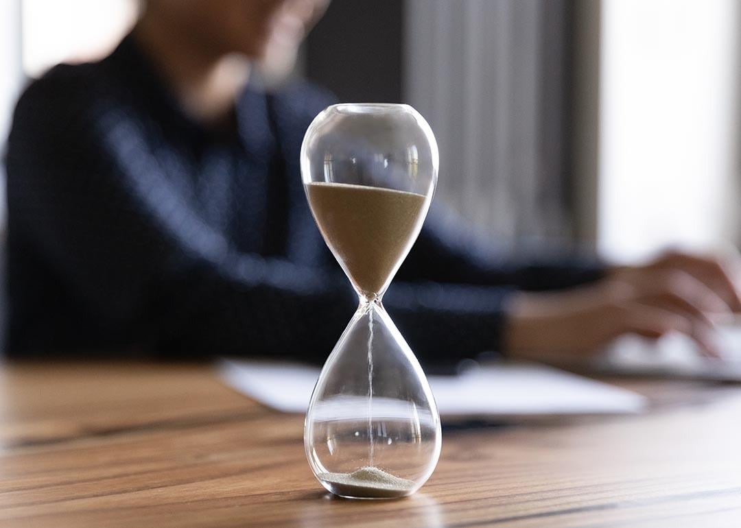 Close up view of hourglass stand on wooden office table measuring time, person working on laptop in the background blurred.
