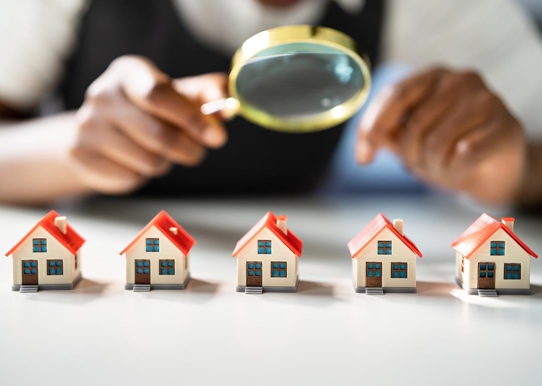 Five miniature houses with red roofs lined up on table, with magnifying glass held above them by a person symbolizing home and neighborhood appraisals.