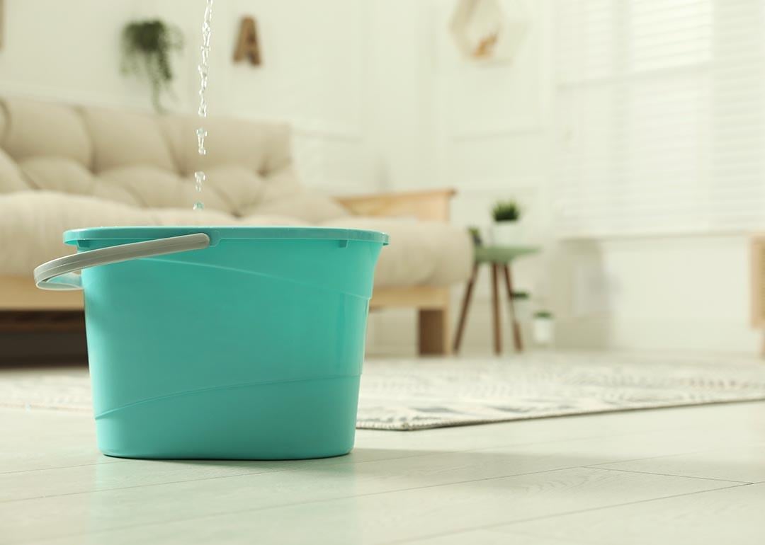 Close up of turquoise-colored plastic bucket on floor in living room with water dripping from ceiling into it.