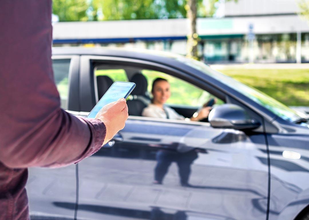 Arm of person in focus in the foreground, holding smartphone, with blurred dark car pulling up in the background, driver visible through the passenger window.