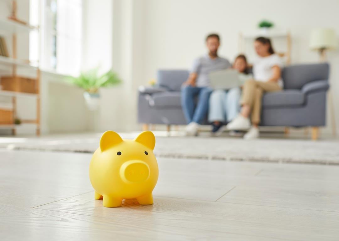 Yellow piggy money box standing on floor in the living room and a parents with a child girl sitting on sofa at home in background.