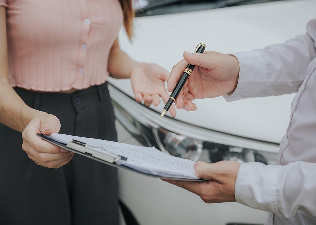 Close up of midsections of two people whose faces are not visible looking at a clipboard with forms on it with a white car in the background.
