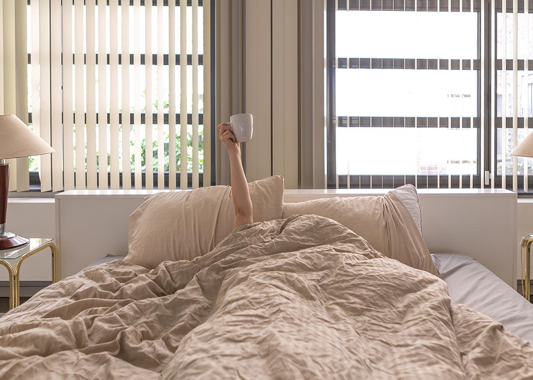 Hand holding a coffee cup extending upward from person whose face and body are under the covers in bed.