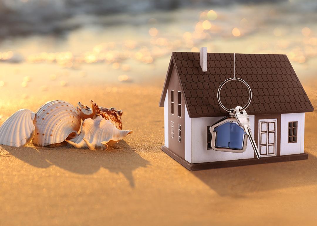 A model of a house with keys on a sandy beach beside seashells.