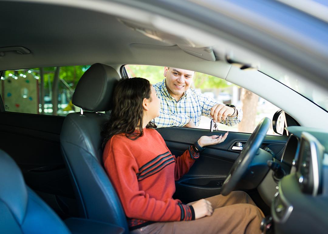 A teenage daughter inside a car receiving its keys from her proud dad.