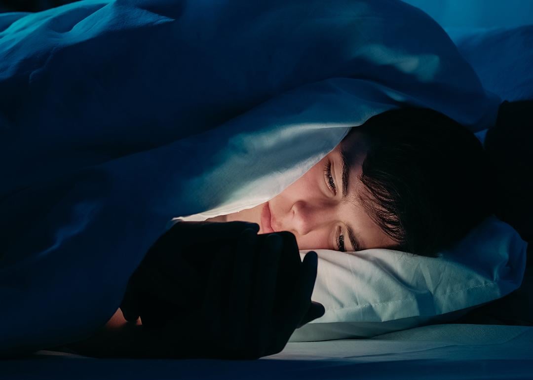 A male teenager using his smartphone under bedsheets at night.