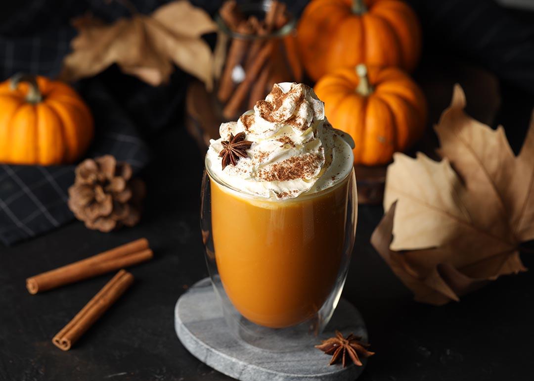Glass of pumpkin spice latte with whipped cream and ingredients like cinnamon sticks and fall decor on black table.