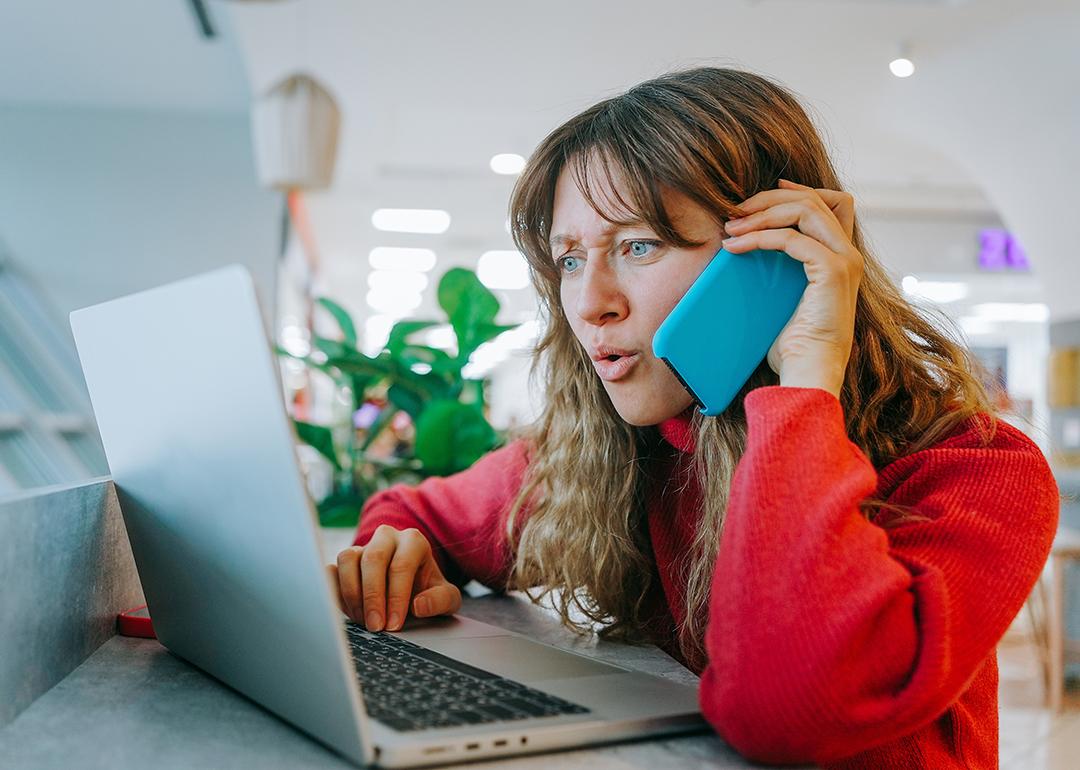A woman talking on her smartphone while also booking flights using her laptop.