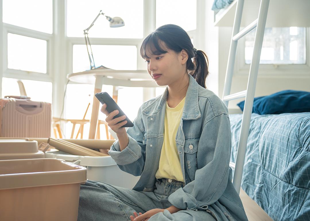 A college student using her smartphone in her dorm while unpacking personal items.
