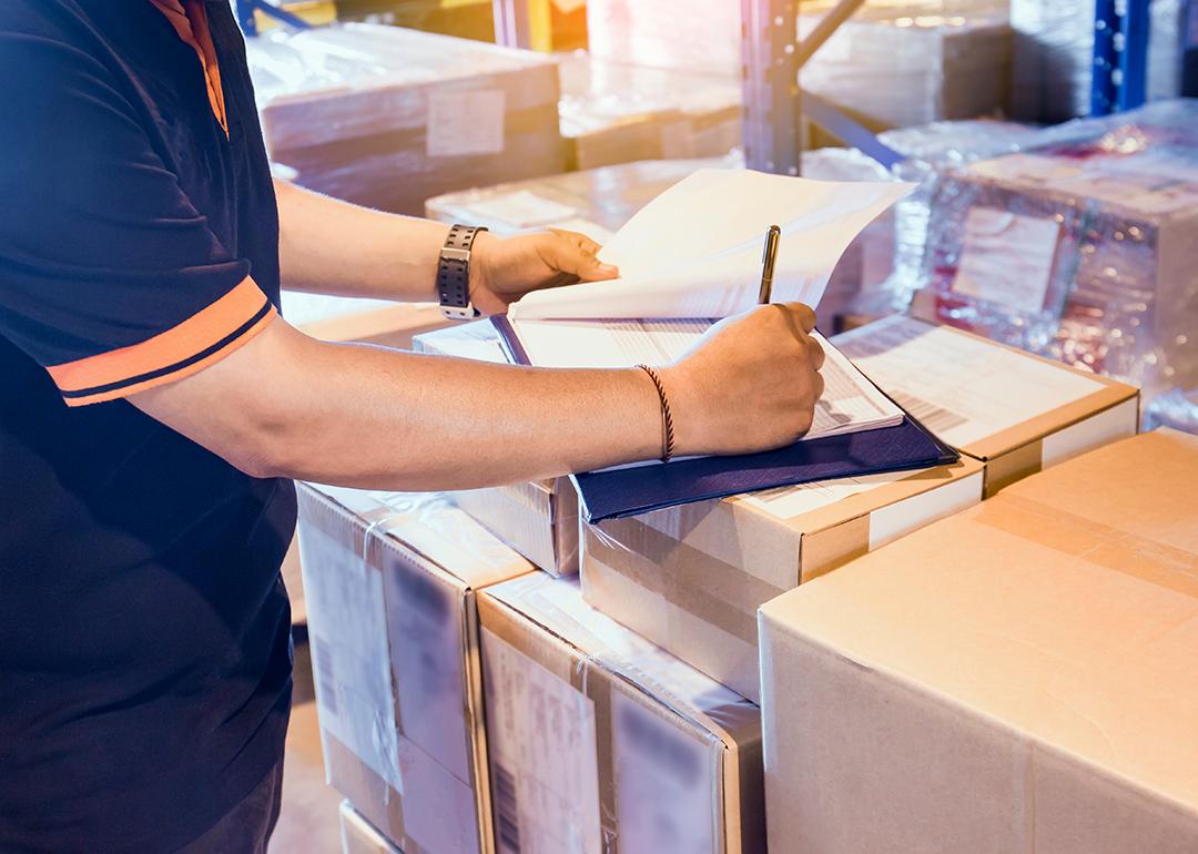 Warehouse worker writing on a clipboard on top of boxes as part of checking inventory.