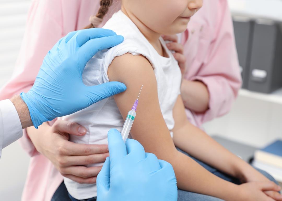 closeup of child's arm with vaccine needle held next to it by a person with blue gloves, an adult in a pink shirt holds child from other side.