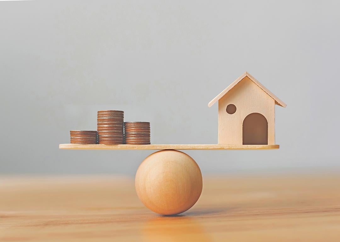 Wooden scale balancing a mini wooden figure of a house on the right and stack of coins on the left.