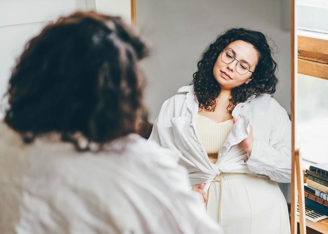 A curvy woman stands in front of a mirror, styling her outfit with a long sleeve shirt.