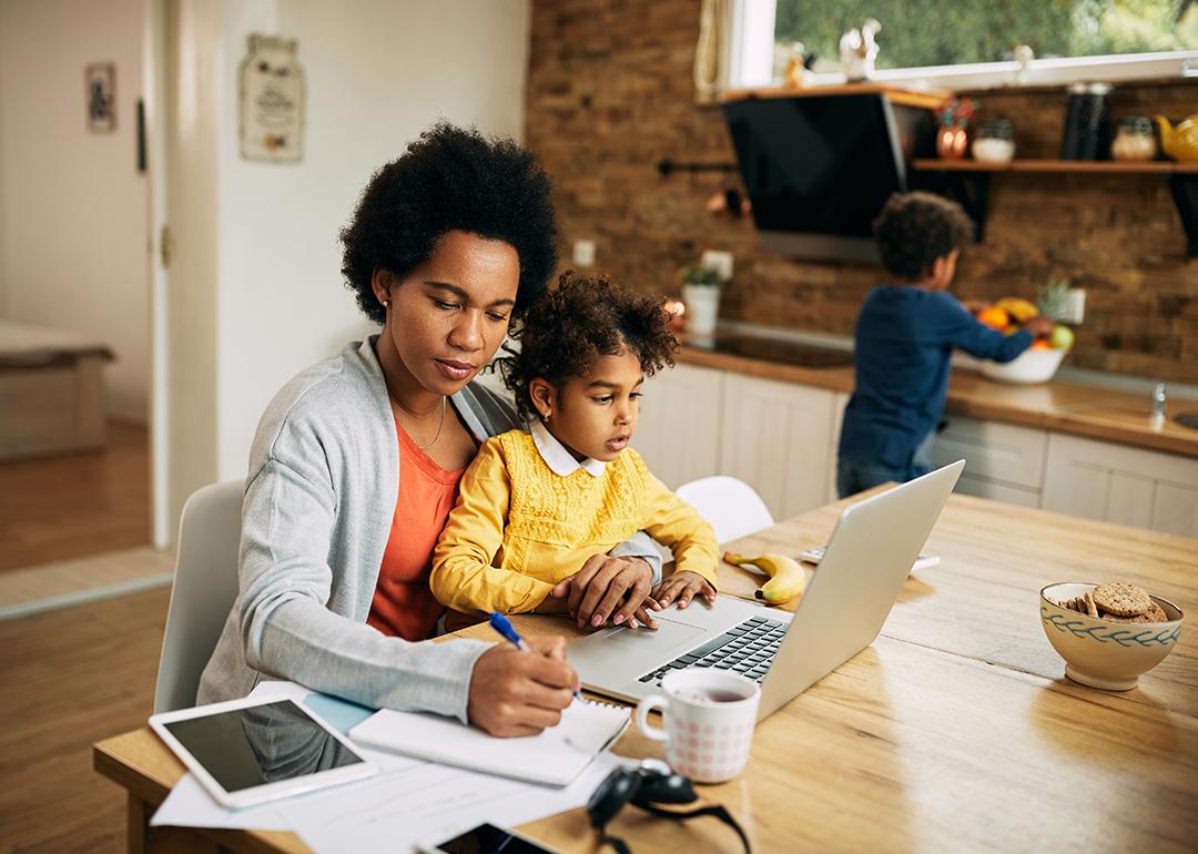 A black mother takes notes as she works at home while her young daughter sits on her lap.