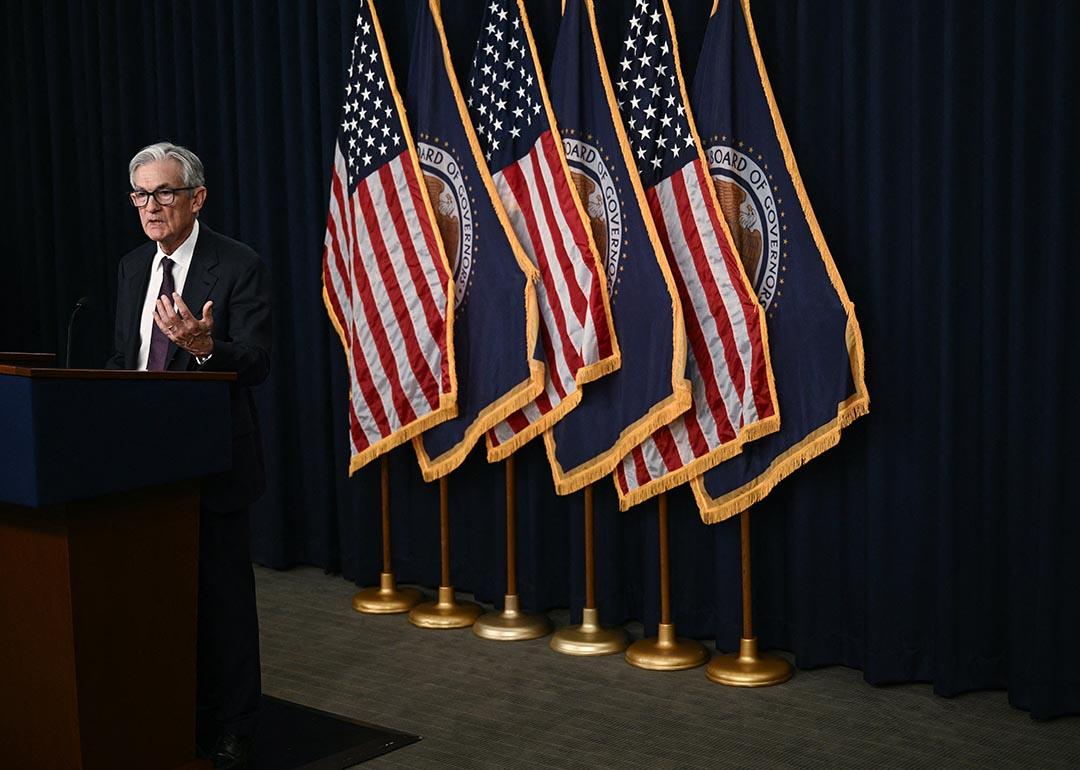 Fed chair Jerome Powell speaking at a podium with six flags behind him, 3 U.S. flags and 3 Board of Governors flags in alternating pattern.