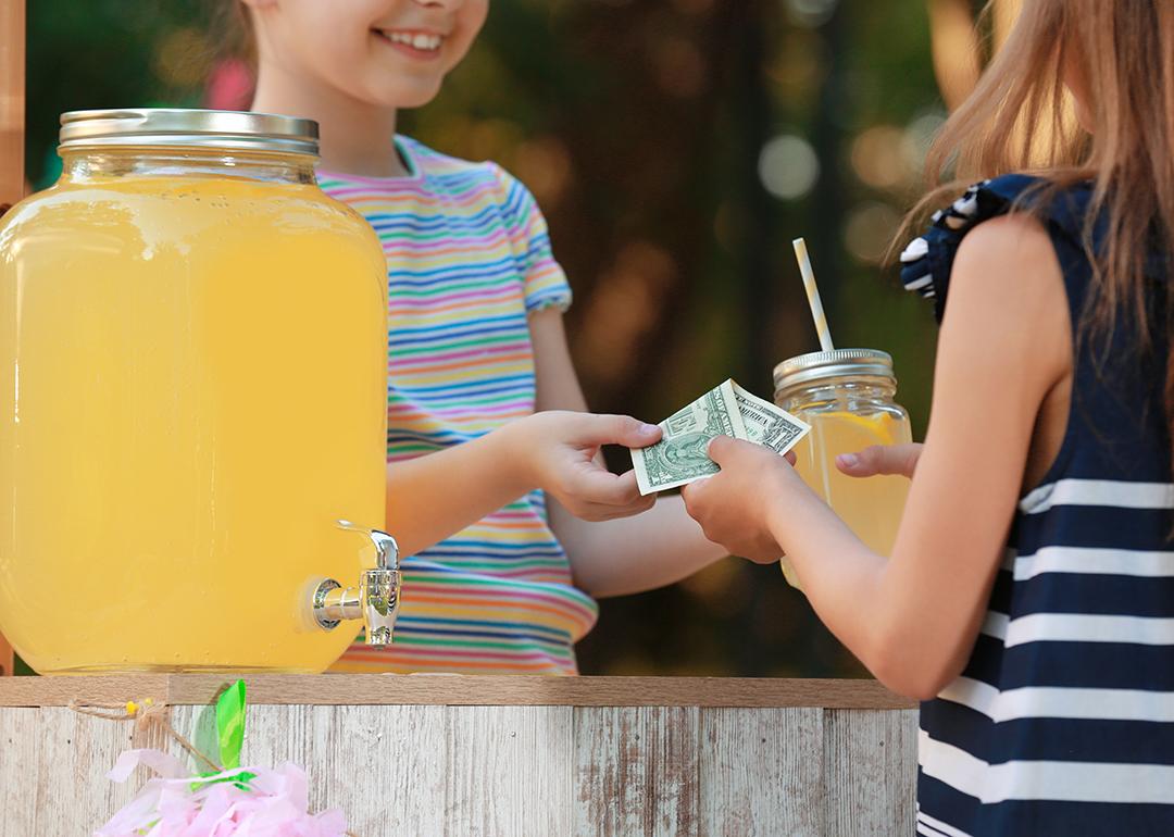A young girl sells lemonade to another young girl at the park.