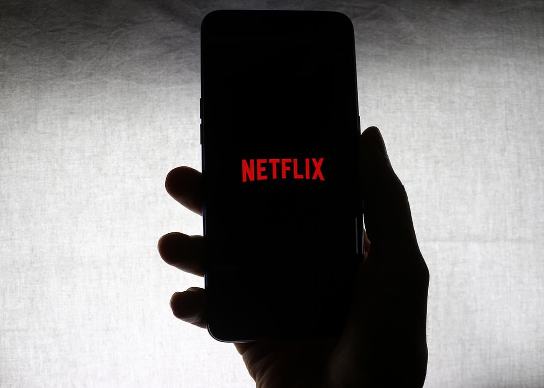 The Netflix logo on the screen of a mobile phone held by a person against a white background.