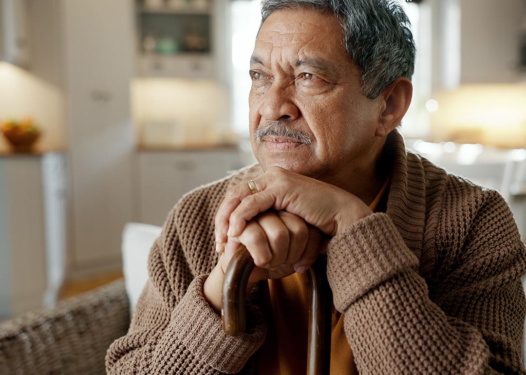 A senior man with a cane thinks deeply while sitting on a sofa.