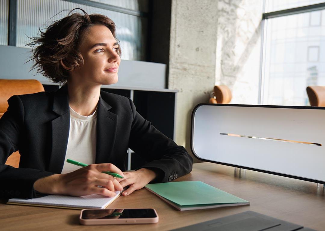 Business woman in office looks toward window thinking while taking notes.