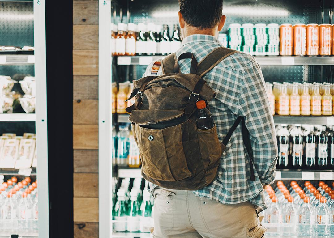 A man traveling with a backpack facing the cold drinks section of an airport food store.