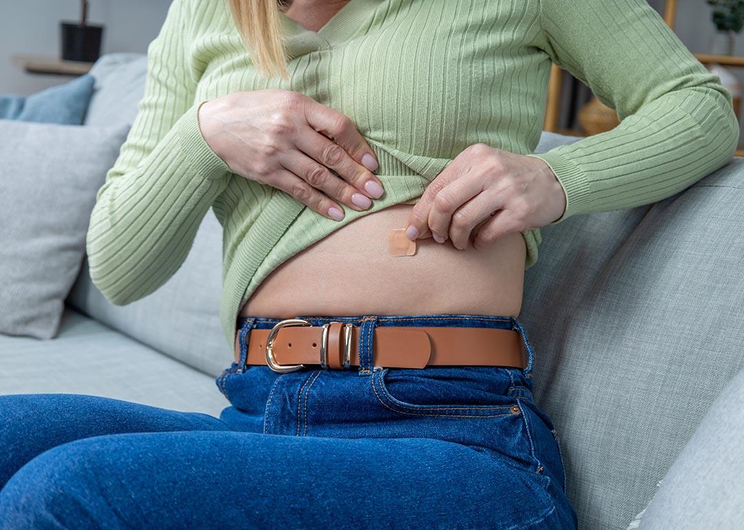 Close up of woman sitting on couch pulling up long sleeve light green shirt to apply hormone replacement patch to her abdomen.