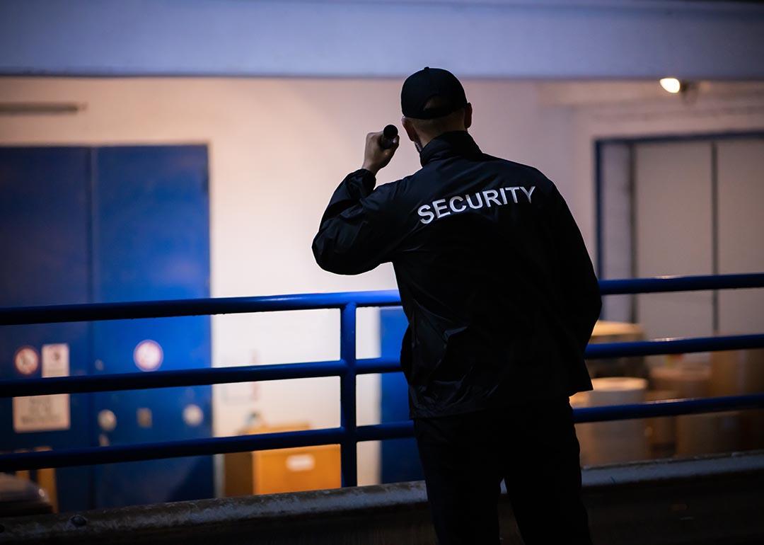 A person standing on a loading ramp outdoors at night wearing dark clothes with the word "SECURITY" displayed on the back of a shirt shines a flashlight toward a white wall with blue doors.