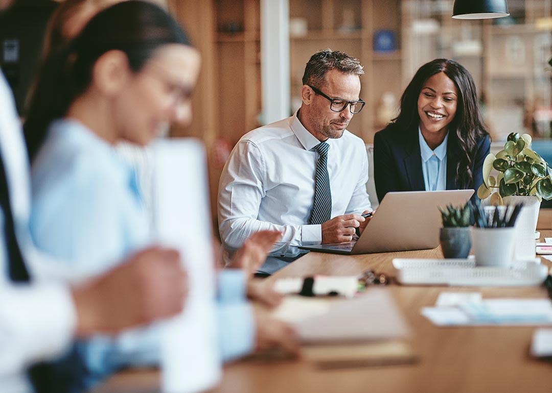 Two diverse businesspeople smiling while working on a laptop together at the end of a boardroom table in an office.