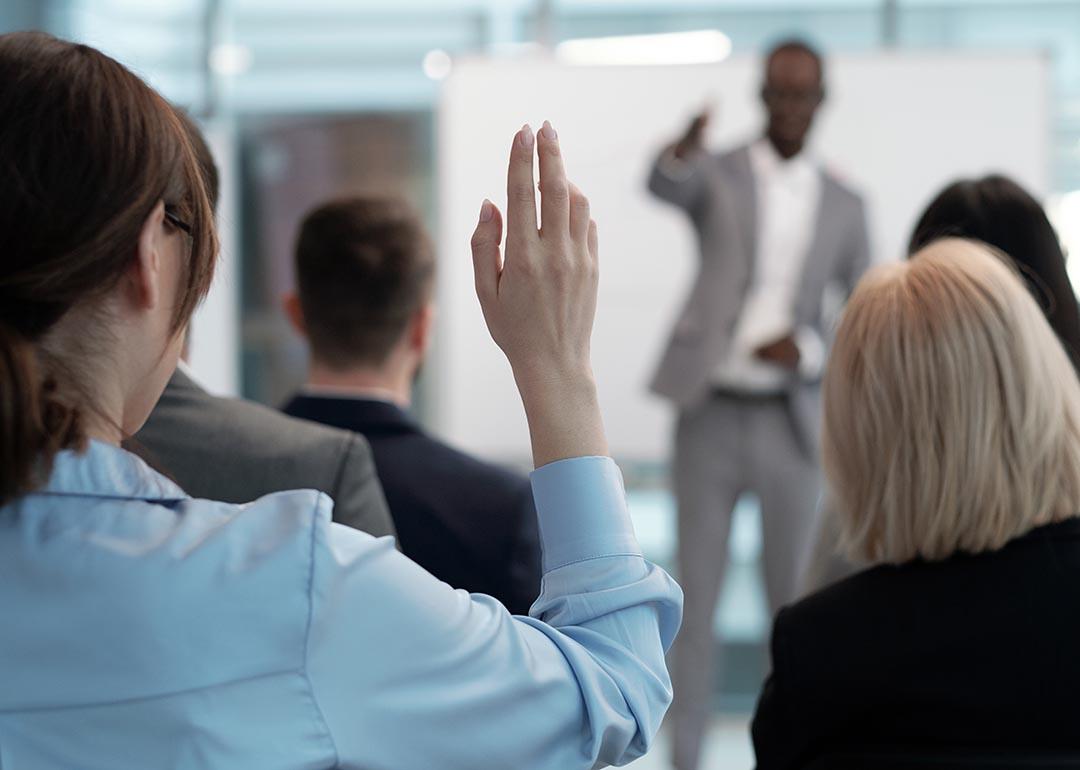 Rear view of raised hand with pointy nails of young employee wishing to answer question of lecturer while sitting behind group of other speakers at conference.