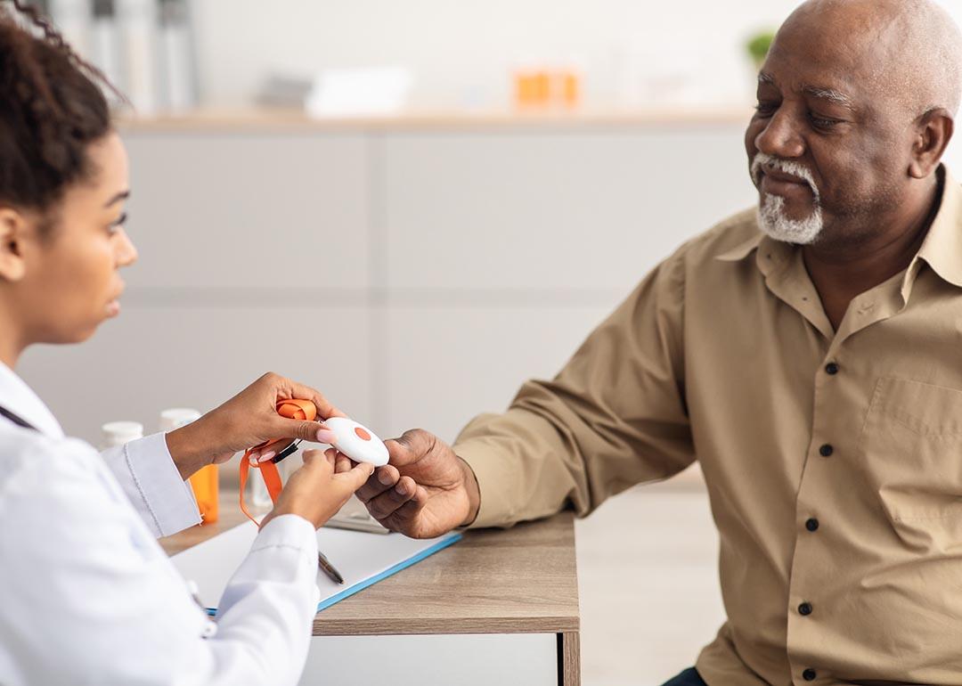 Portrait of black medical worker holding personal alarm button and giving it to mature patient, sitting at table in office, explaining how to use medical alert.