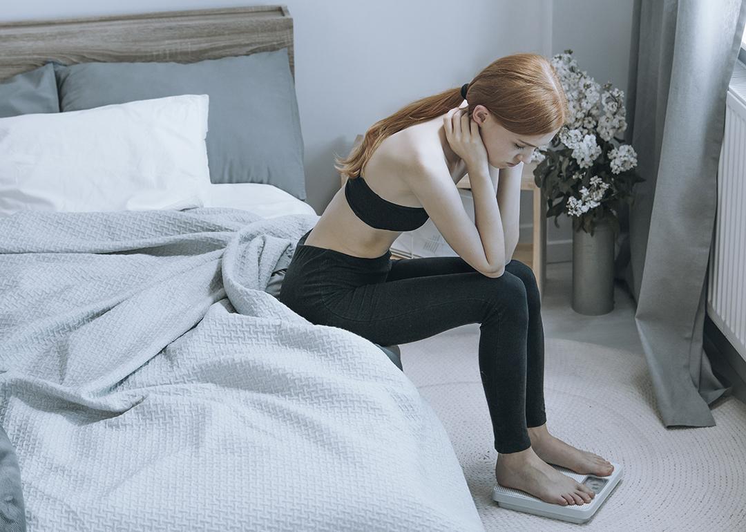 A young woman sits by her bed while pensively checking weight on a scale.