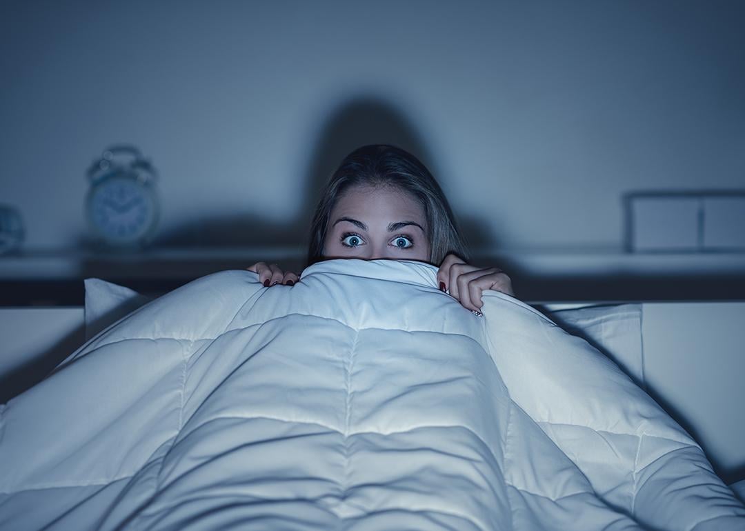 Stylized photo of woman hiding under covers with wide eyes, illuminated by screen from TV.