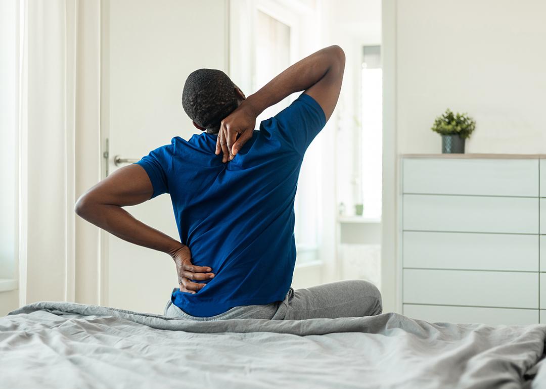 A young black man stretches and reaches for his back after getting up from bed.