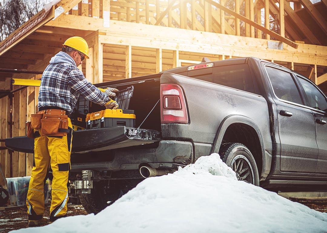 A construction worker in yellow gear prepares tools from a truck at a snowy building site with wooden building framing in the background.