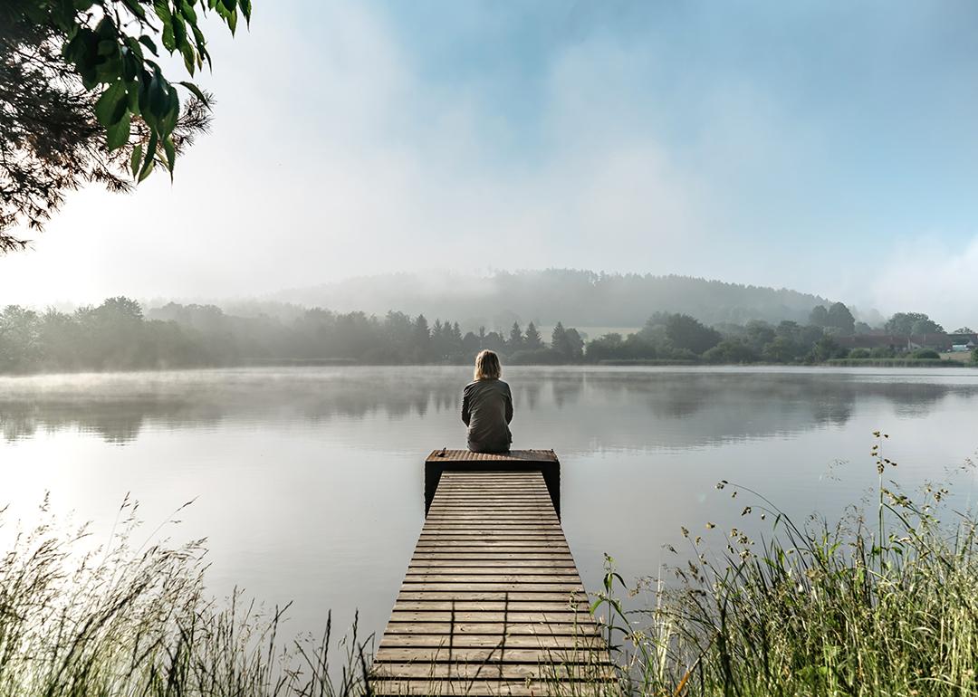 A woman sits quietly at the end of a small pier by a lake during a calm, misty morning.
