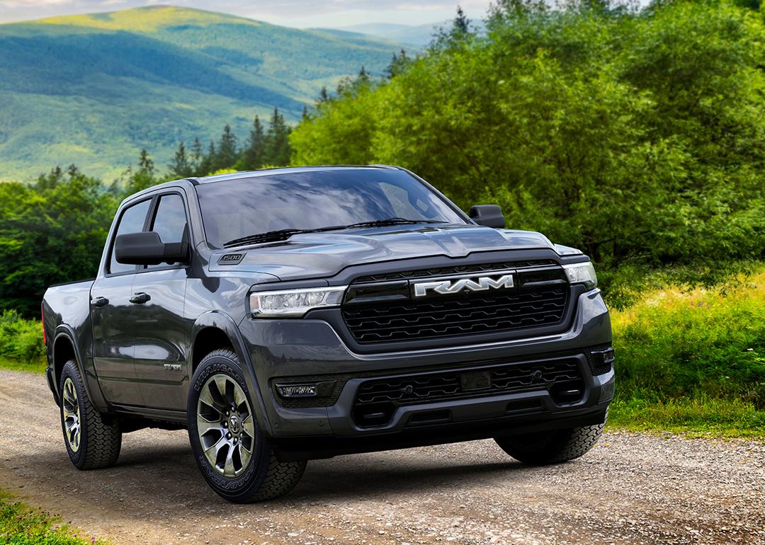 Black 2026 Ram 1500 REV Bighorn driving up dirt road with rolling green hills in background.