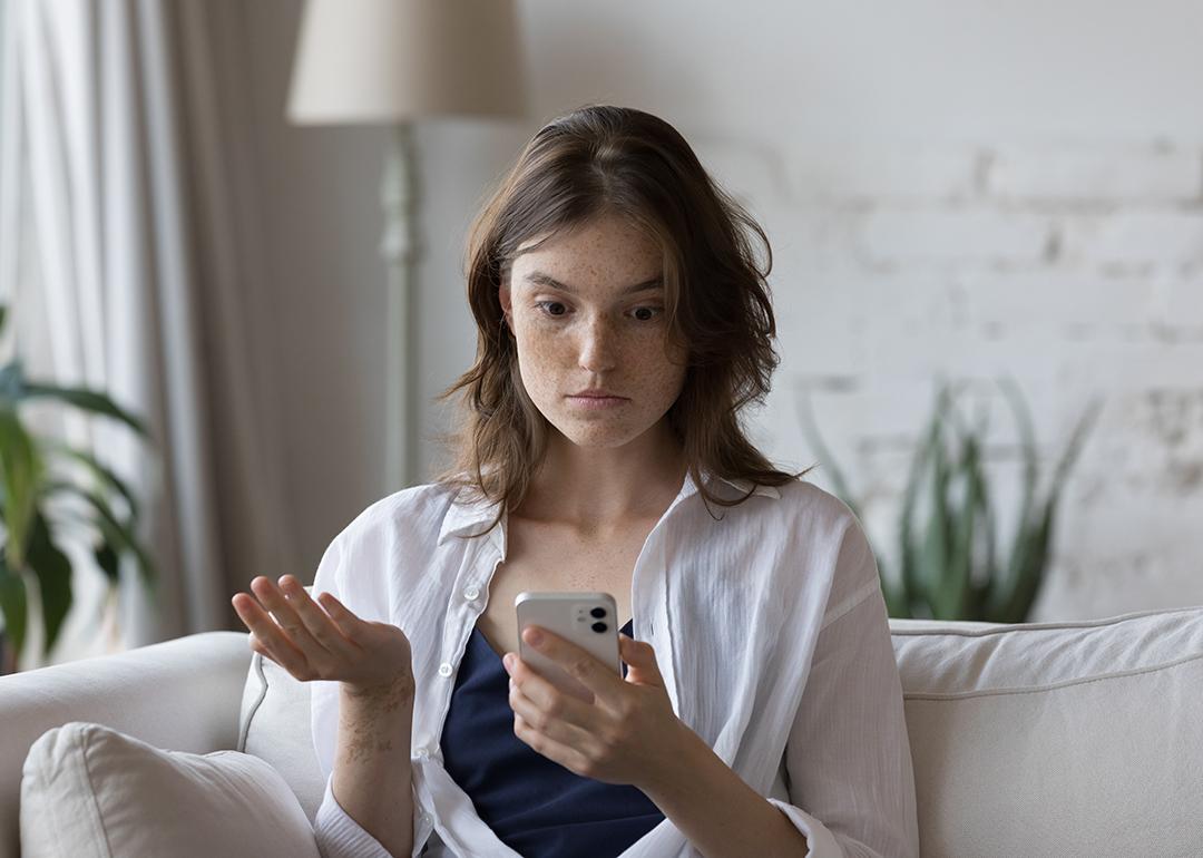 A woman sitting on a couch at home looking concerned at her phone.